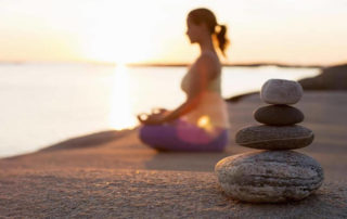 Woman meditating on beach with little stacked pebbles on the foreground.