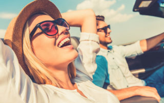 Woman laughing with her hands behind her head driving with a man.